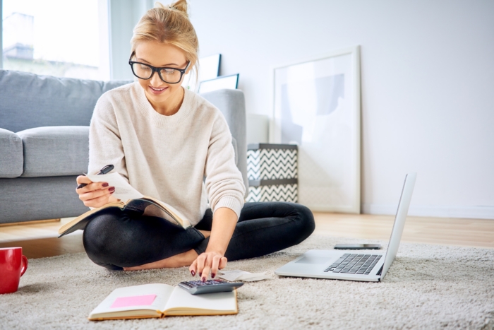 Young millennial woman sitting on the living room floor using a calculator and laptop to plan her budget.