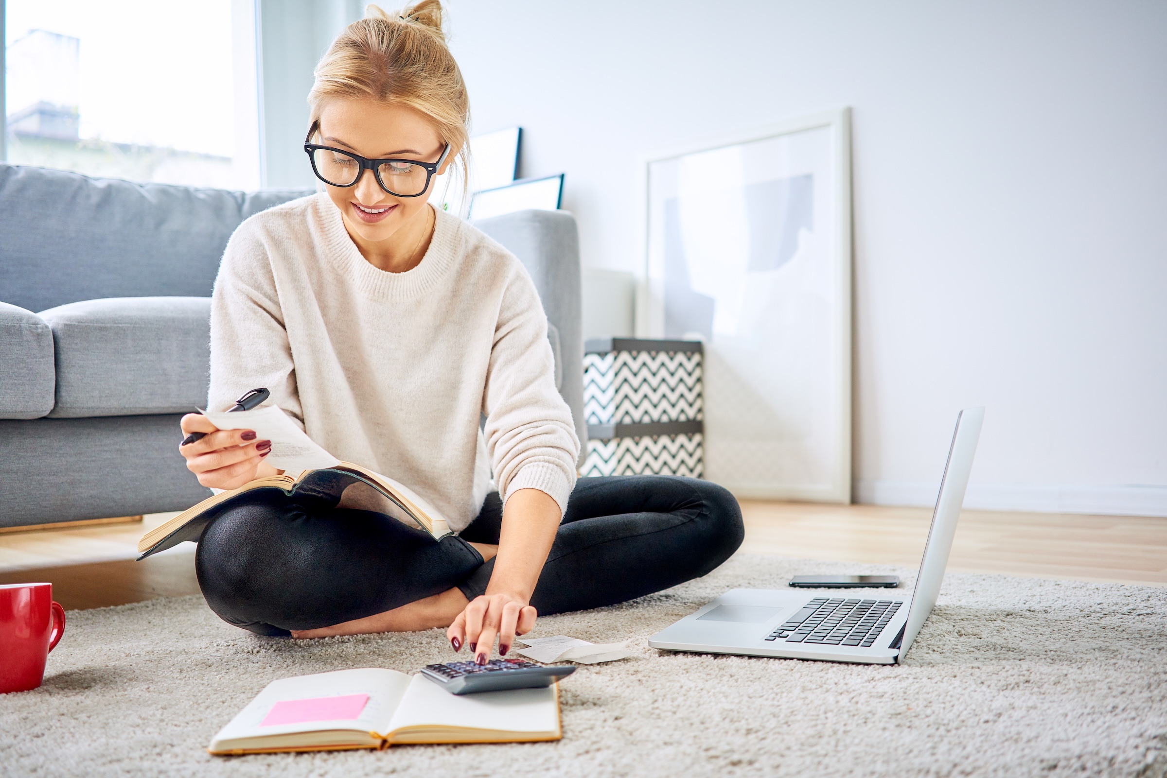 Young millennial woman sitting on the living room floor using a calculator and laptop to plan her budget.